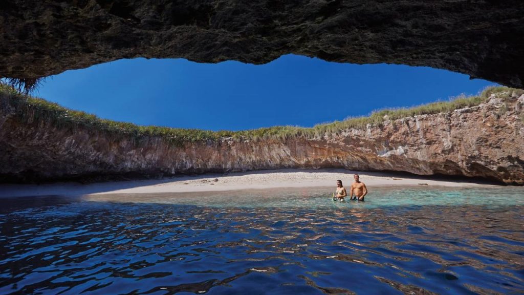A couple wade through the shallows by a beach hidden within a cave