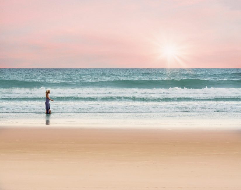 woman in thought walking along a beach, with golden sand, blue sea and a pink early sunset