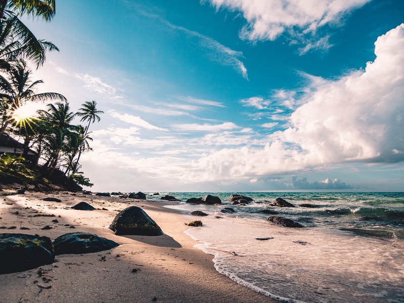 An expansive blue horizon zooms off into the distance away from a rattan recliner with a parasol on a huge white sandy beach