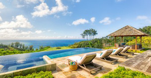 Four sun beds lined up along a pool set among insanely green foliage looking across the see to where the sun is going to set in Hawaii